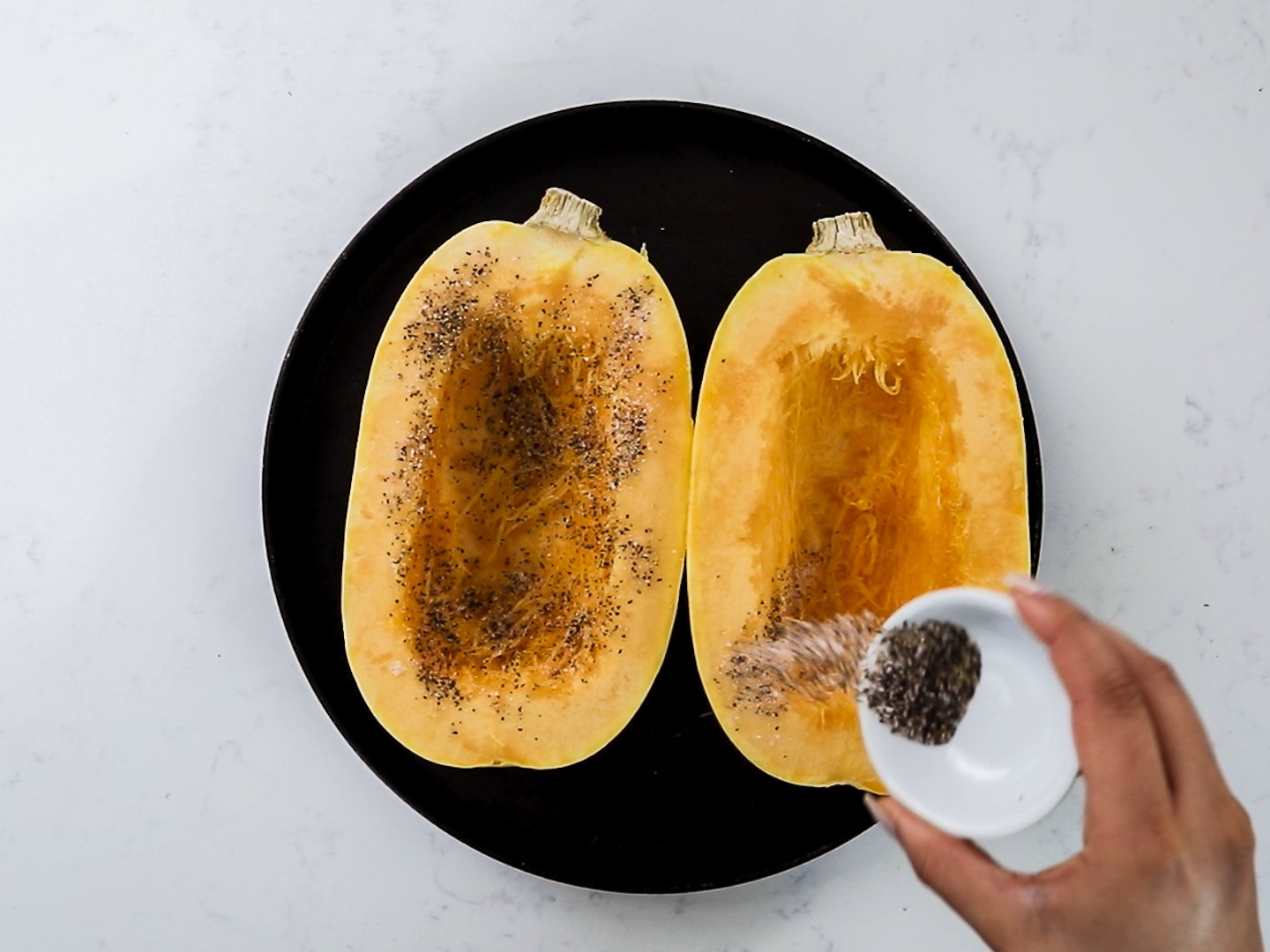 A hand sprinkling a mixture of salt and pepper in a ramekin over two halves of spaghetti squash.