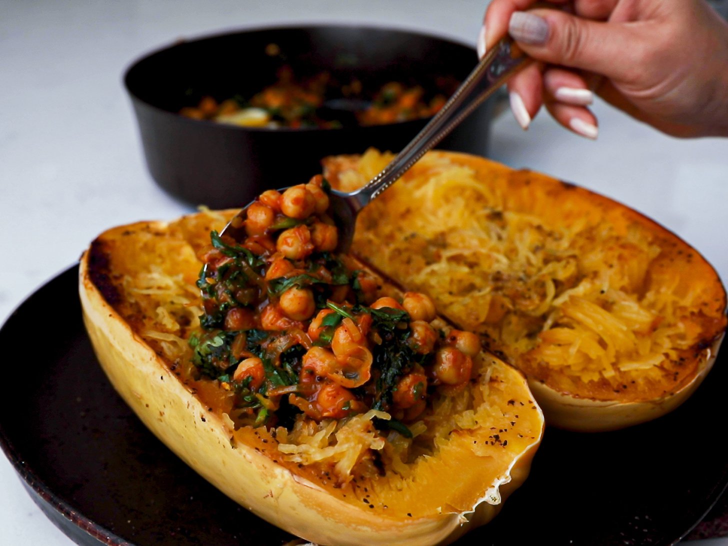 A hand holding a spoonful of chickpea and greens curry to stuff a cooked spaghetti squash half.