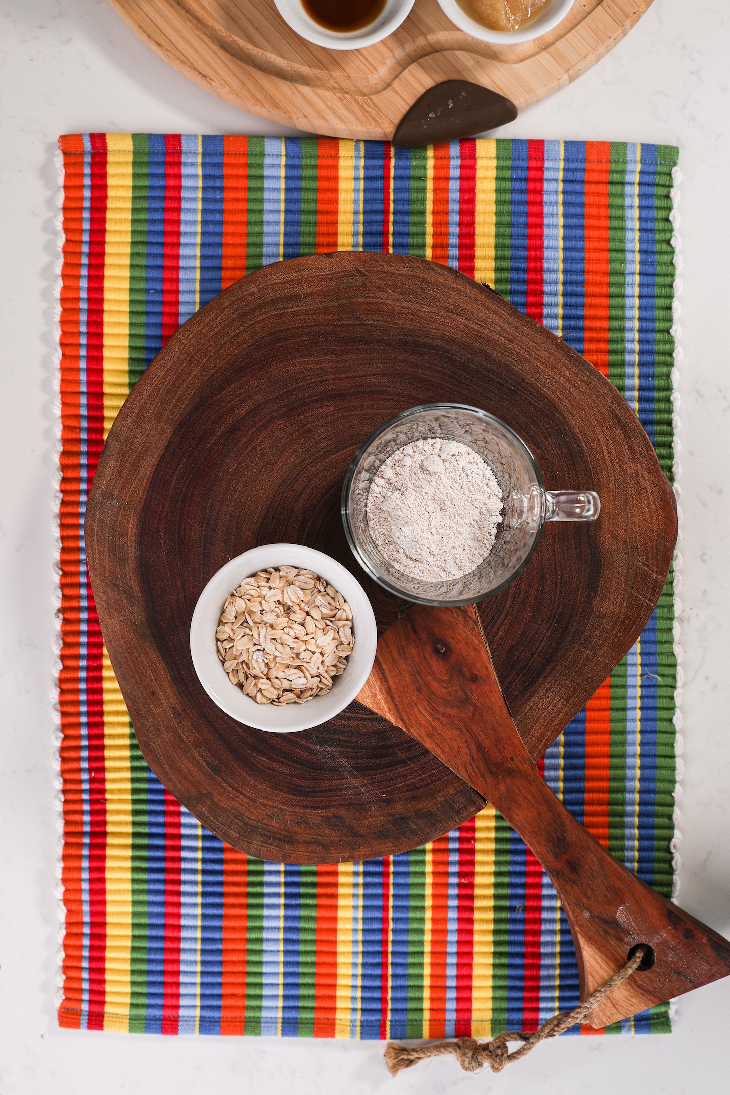 A bowl of rolled oats and a mug of flour on a wooden board that's placed on a colourful place mat.