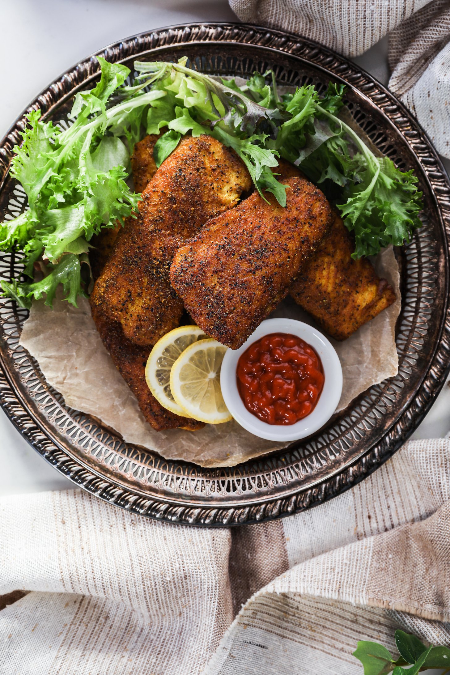 Flat lay image of a pile of crispy fillets with lettuce, red sauce and lemon slices on a tray.