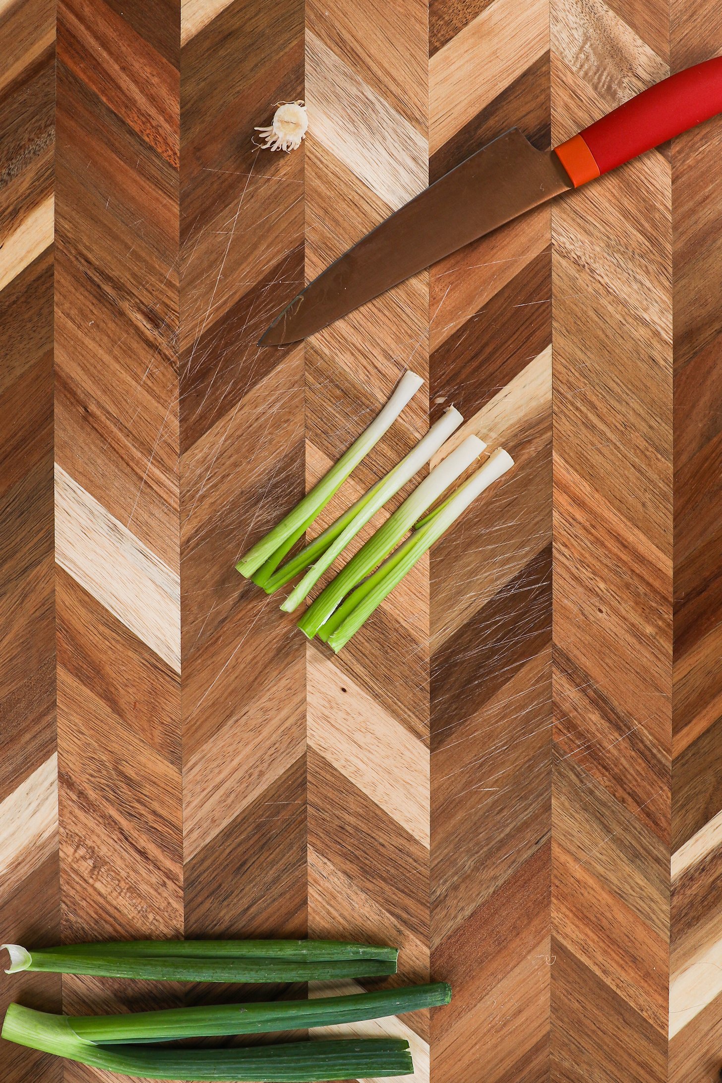 Four strips of spring onion with a red knife on a checkered wooden board.