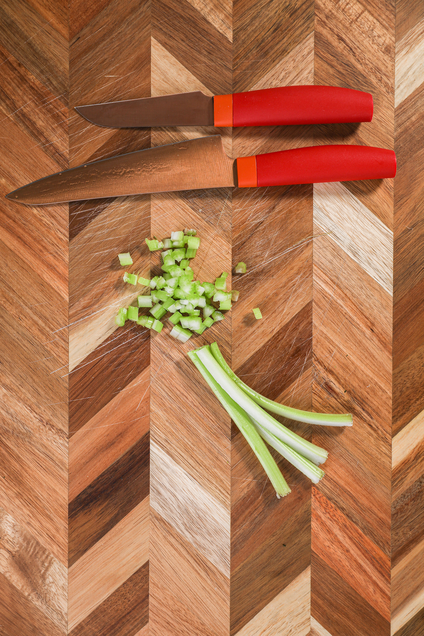 Four strips of celery and finely chopped celery pieces with two knives on a checkered wooden board.