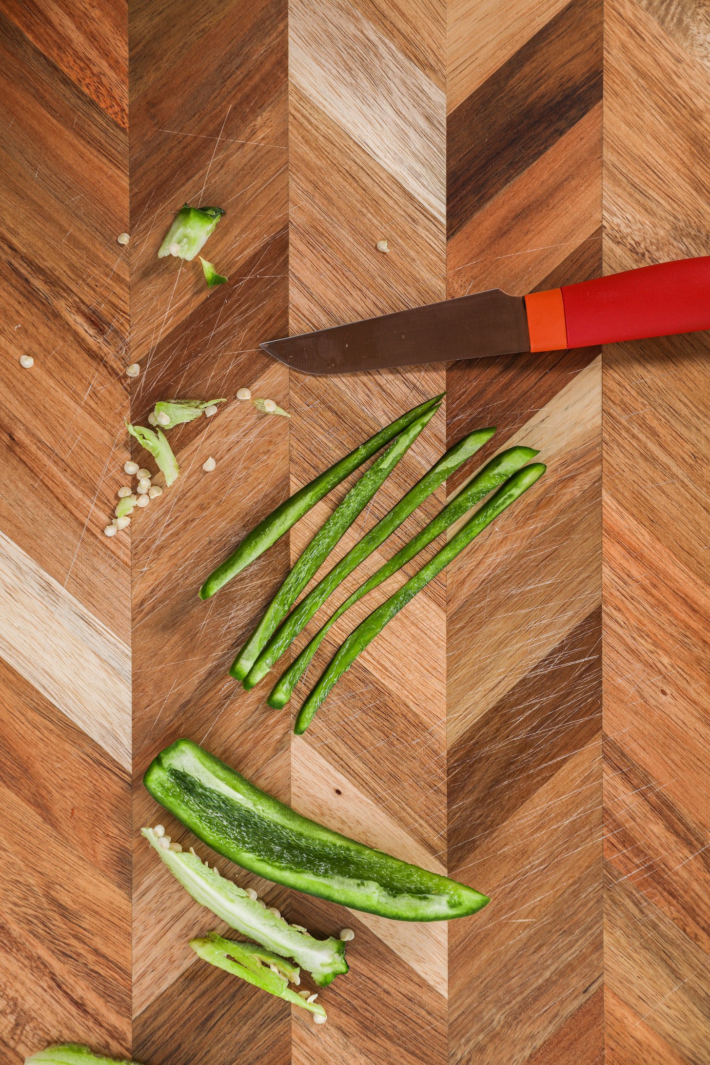 Thin strips of green chilli pepper with a red knife on a checkered wooden board.