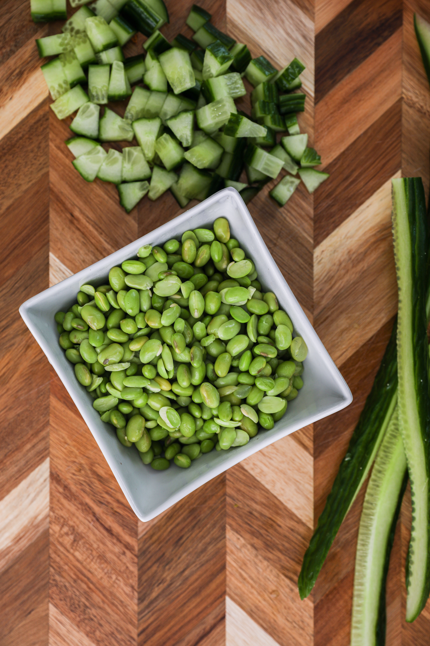 A bowl of edamame and chopped cucumber on a wooden board.