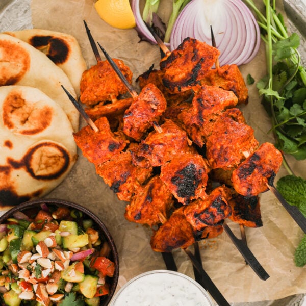 A close up of a pile pf chicken skewers with naan, salad and dip on a round tray.
