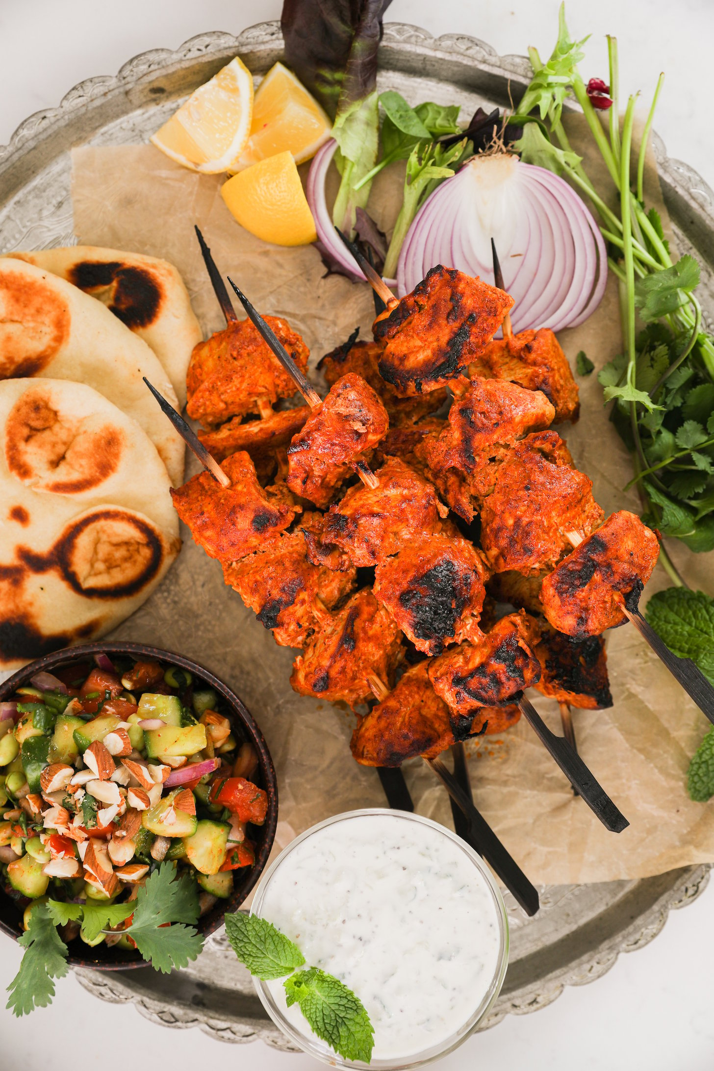 A close up of a pile pf chicken skewers with naan, salad and dip on a round tray.