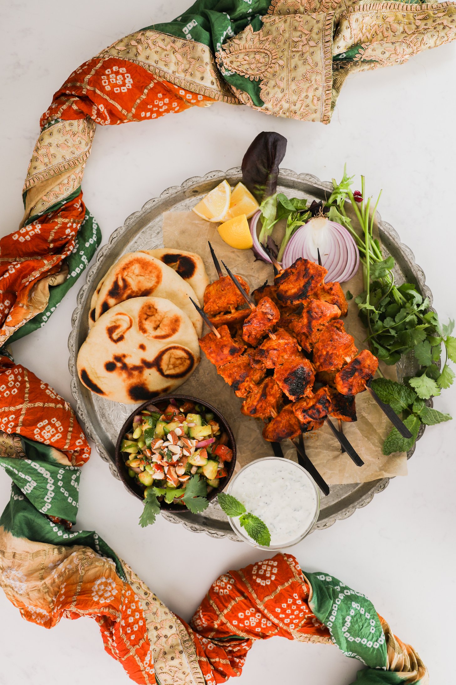 A pile pf chicken skewers with naan, salad and dip on a round tray, styled with a decorative scarf.