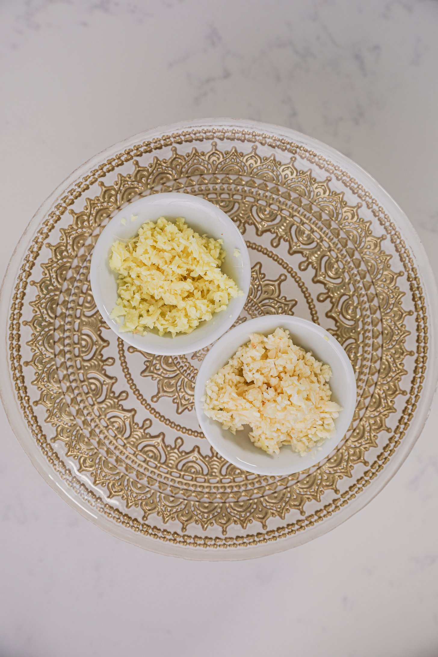 Two ramekins of minced garlic and ginger on a patterned plate.