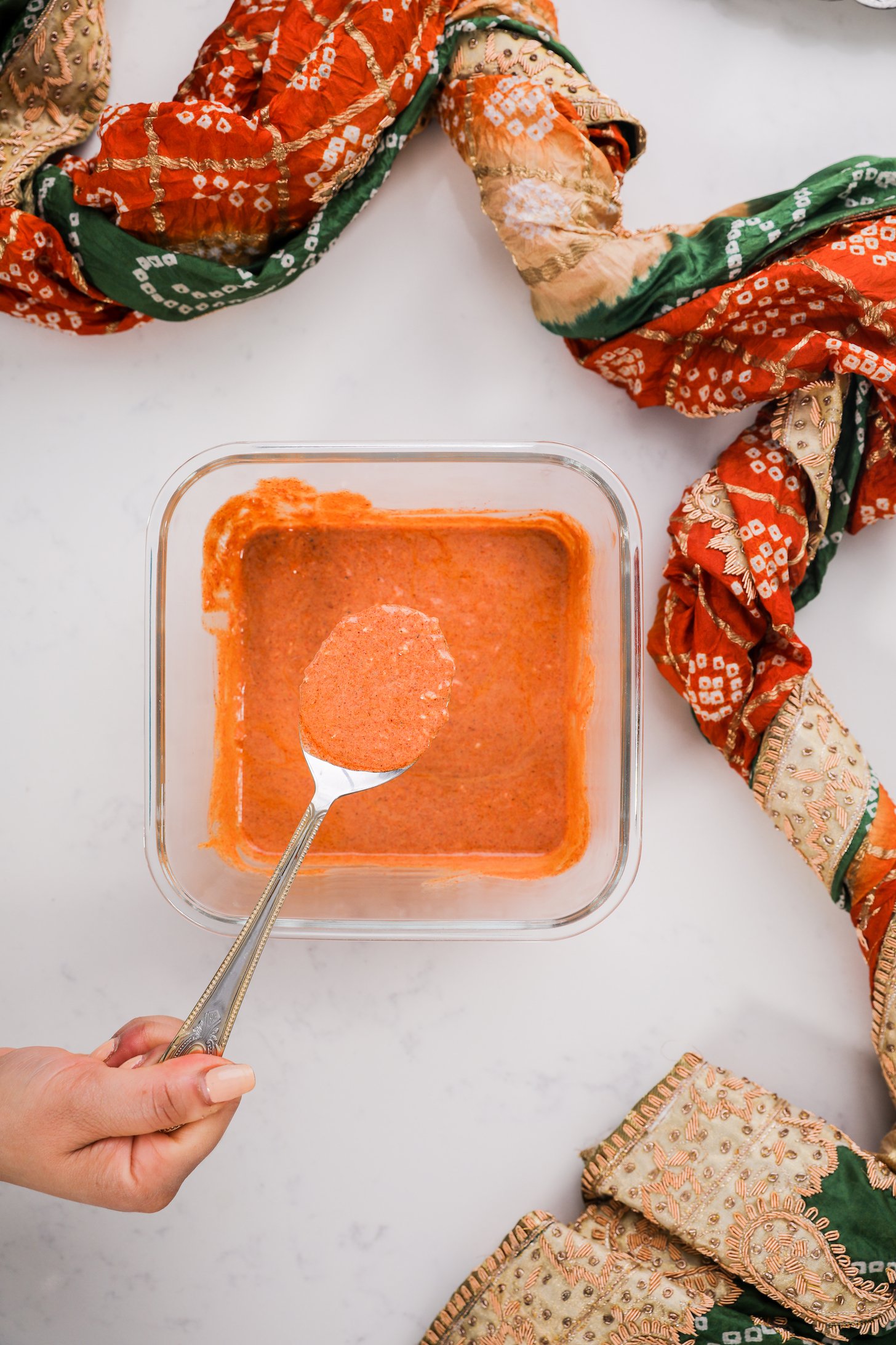 A hand holding a spoon of orange sauce over a bowl of sauce styled around a decorative scarf.