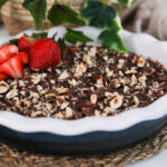 Perspective shot of a bowl of chocolate dessert topped with nuts and some strawberries with a plant in the background.