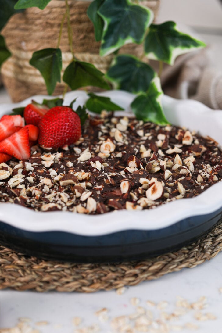 Perspective shot of a bowl of chocolate dessert topped with nuts and some strawberries with a plant in the background.