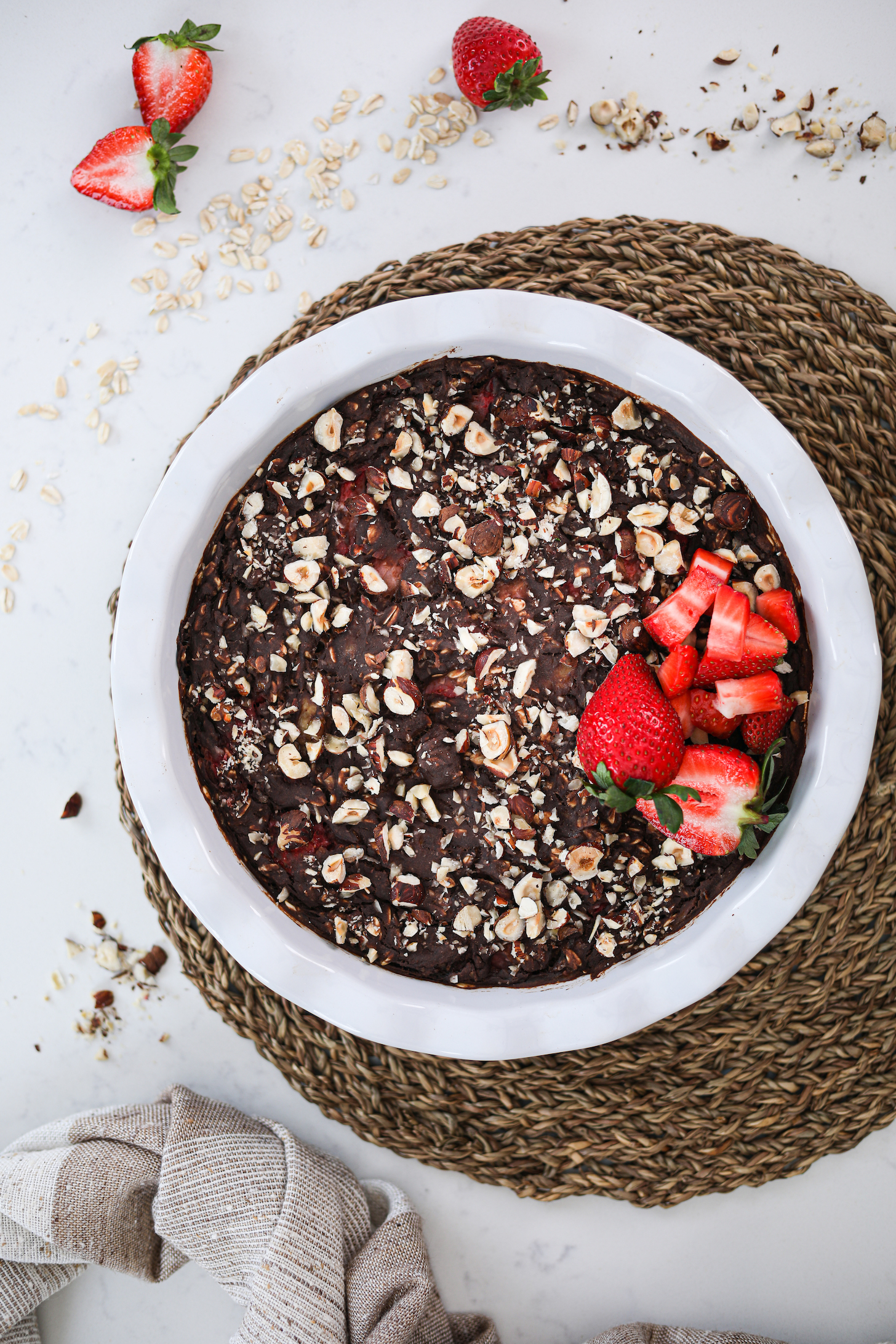 Top view of a bowl of chocolate dessert topped with crushed nuts and a small pile of strawberries styled on a straw mat.