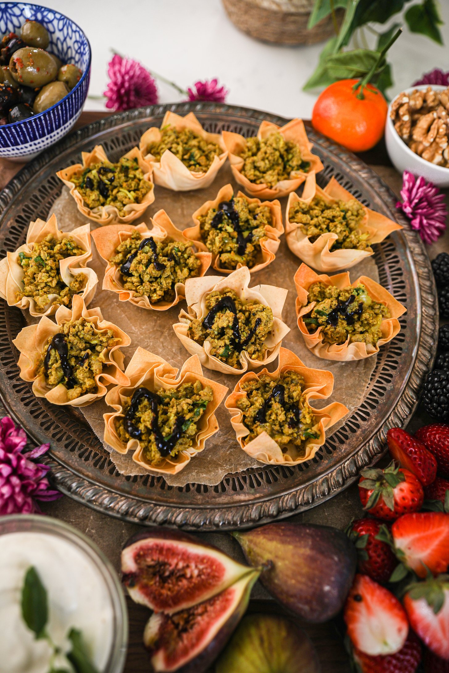 A tray of baked pastry shells filled with minced chicken and topped with a drizzle of a dark dressing surrounded by fruits and nuts.
