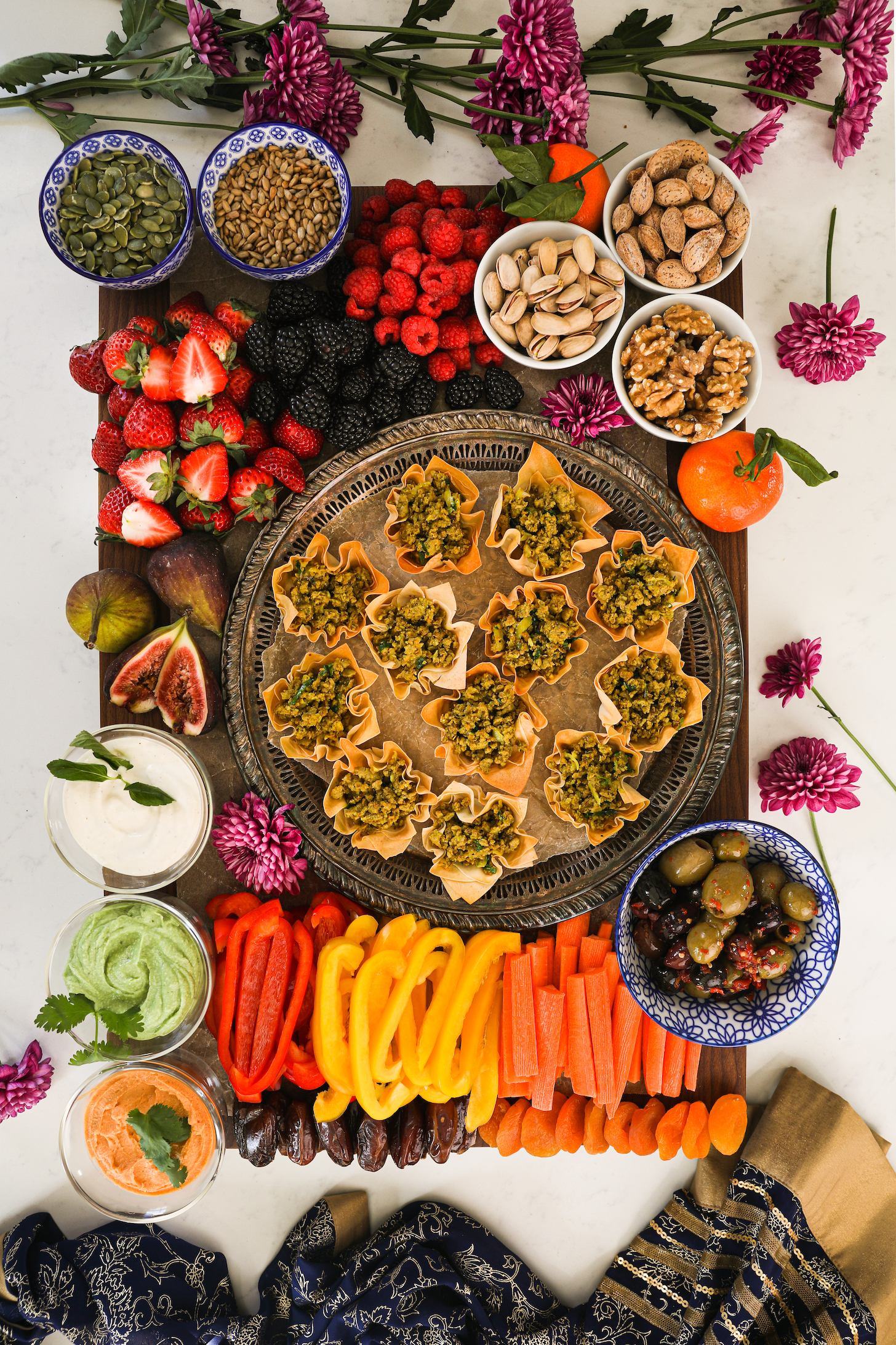 A beautiful and colourful flatlay display of food with a tray of baked pastry shells filled with chicken, fruits, veggies, nuts, olives, seeds and dips. Styled with pink flowers and a traditional scarf.