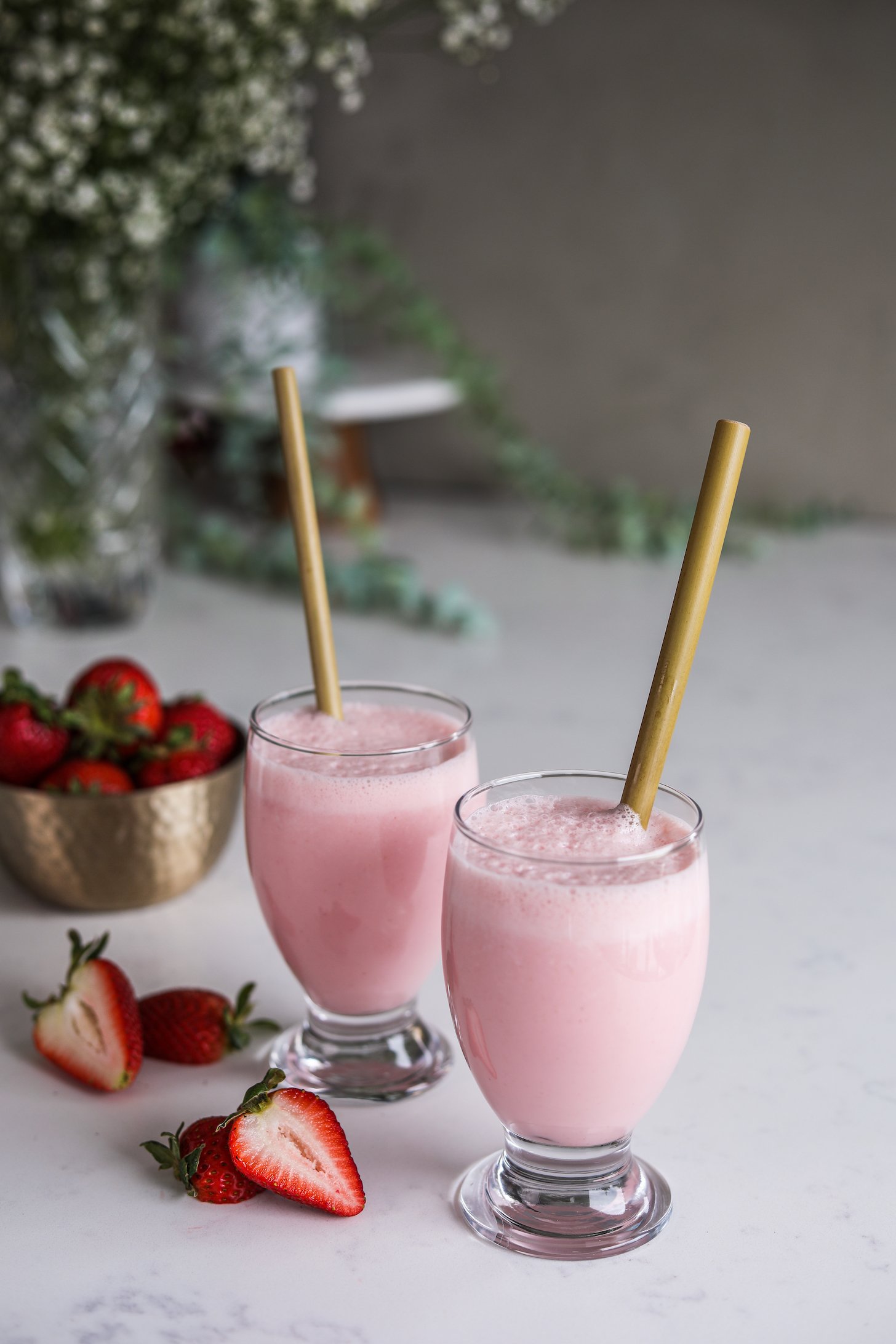 Angled shot of two glasses of a pink frothy drink - each with a straw surrounded by strawberries and plants in the background.