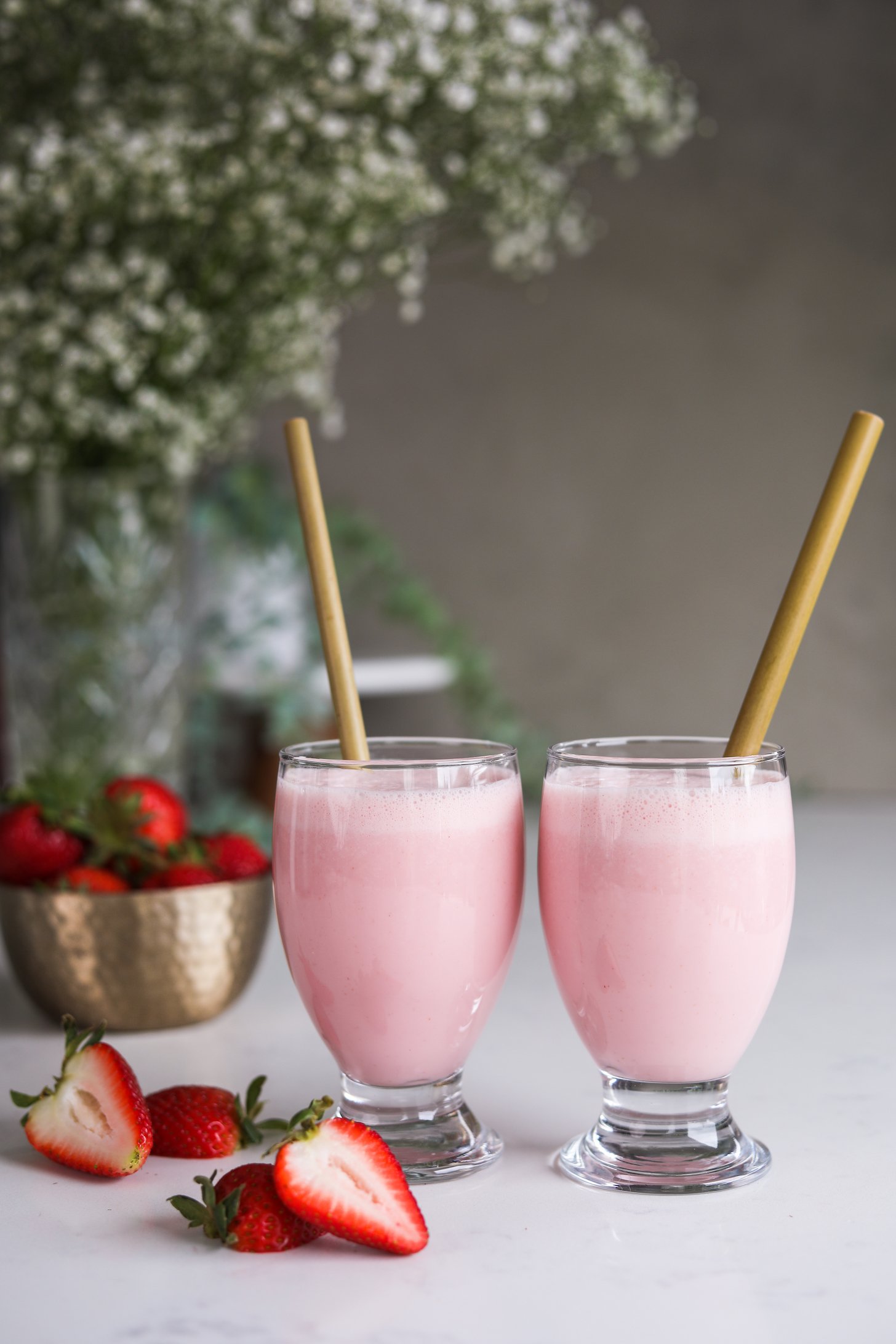 Two glasses of a pink drink - each with a straw with a background of white flowers and fresh strawberries.