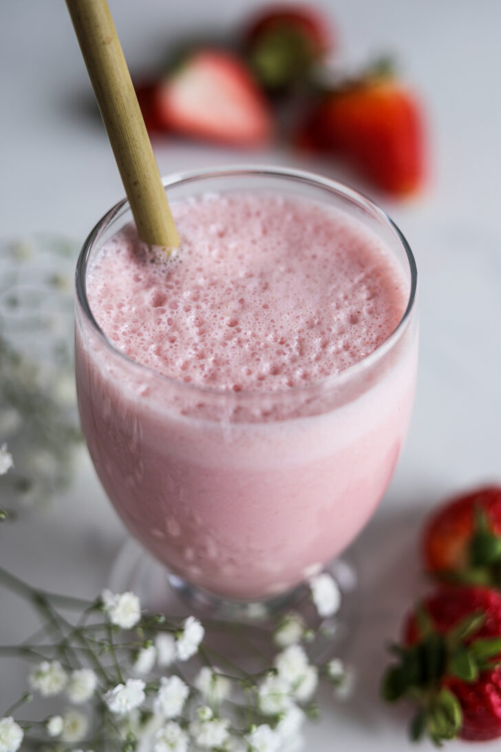A pink frothy drink in a glass with a straw with strawberries in the background.