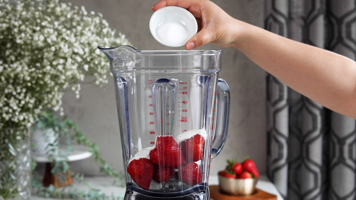 A hand holding a tilted ramekin of sweetener over a blender with berries and yogurt.