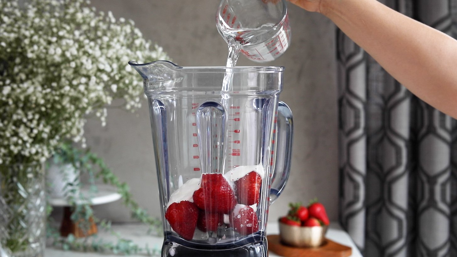 A hand pouring a jug of water into a blender with berries and yogurt.