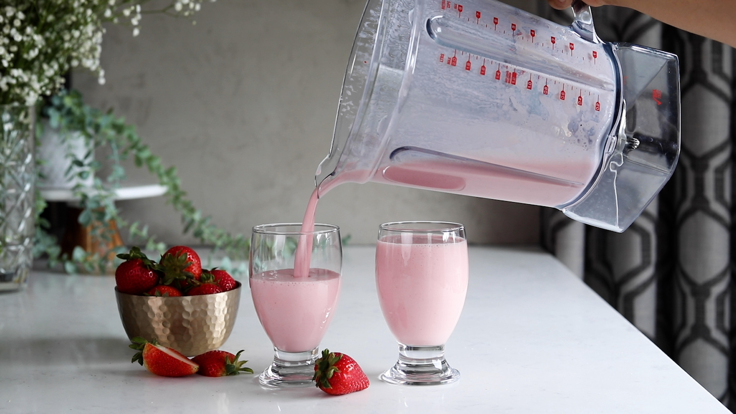 Pouring a pink drink from a blender into two glasses with a bowl of strawberries nearby.