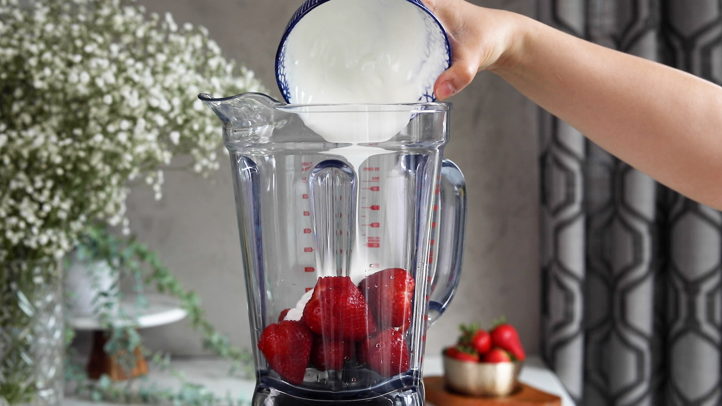 A hand holding a bowl of yogurt and pouring it into a blender with berries.