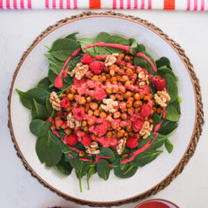 Flat lay of a plate of chickpeas on a bed of spinach topped with walnuts, raspberries and a red dressing with flowers and a kitchen towel styled around it.