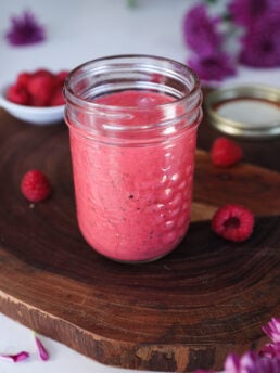 A mason jar of red sauce or dressing on a wooden board surrounded by scattered raspberries and flowers.