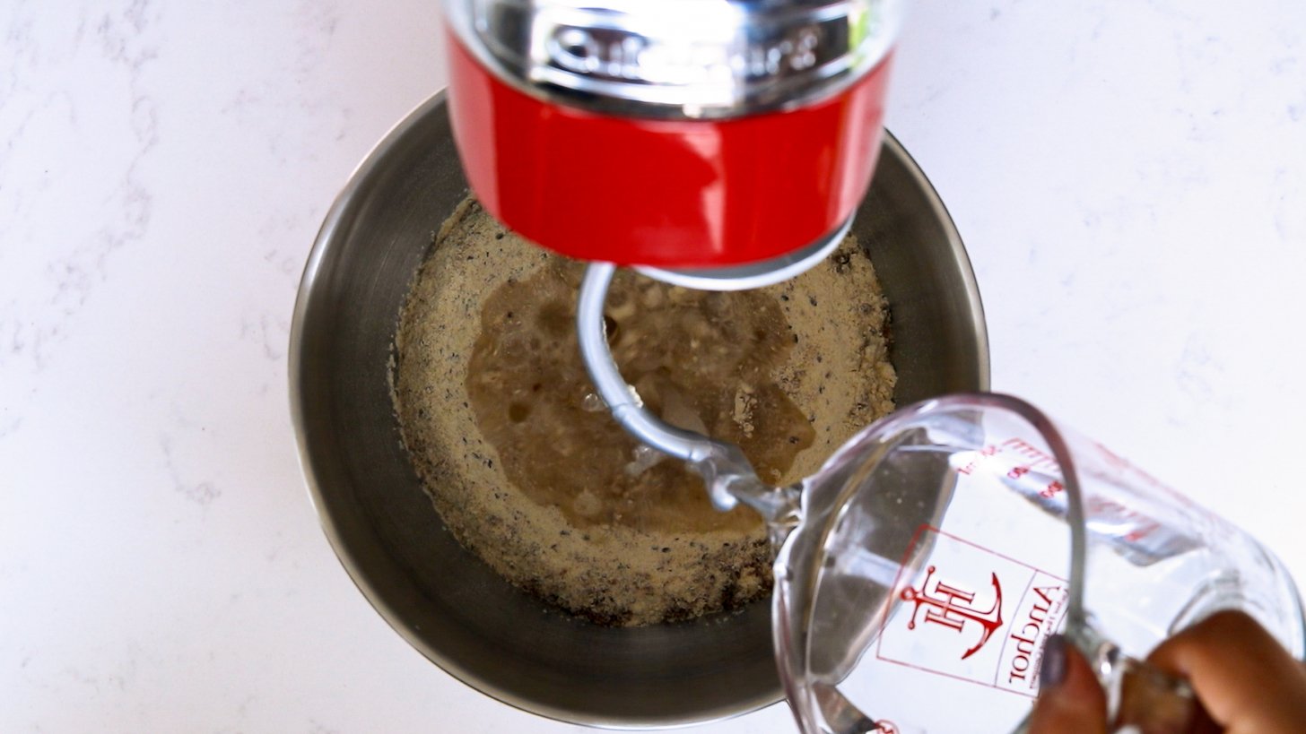 A hand holding a jug pouring water into a stand mixer bowl.