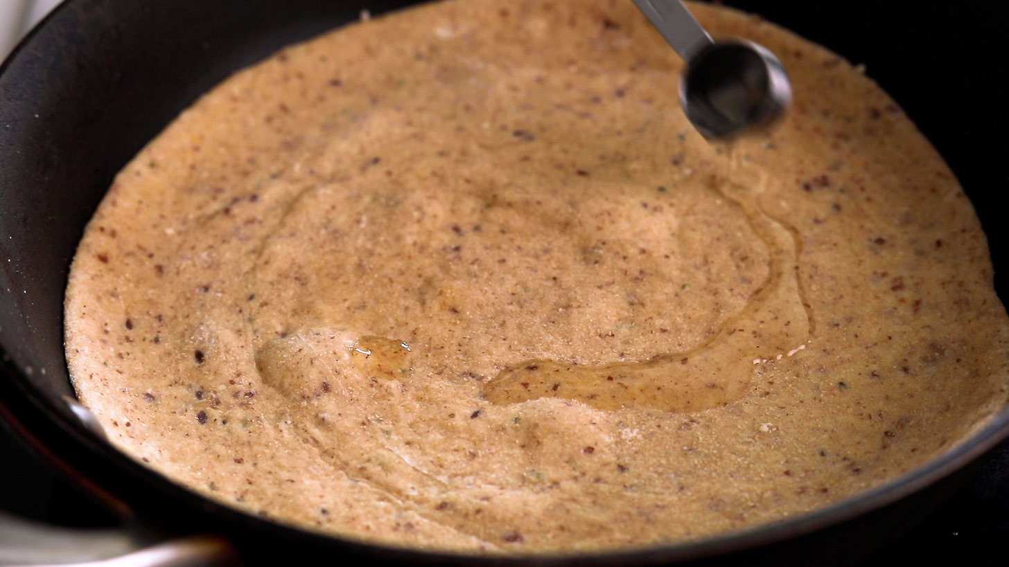 A spoon of oil added to uncooked flatbread in a pan.