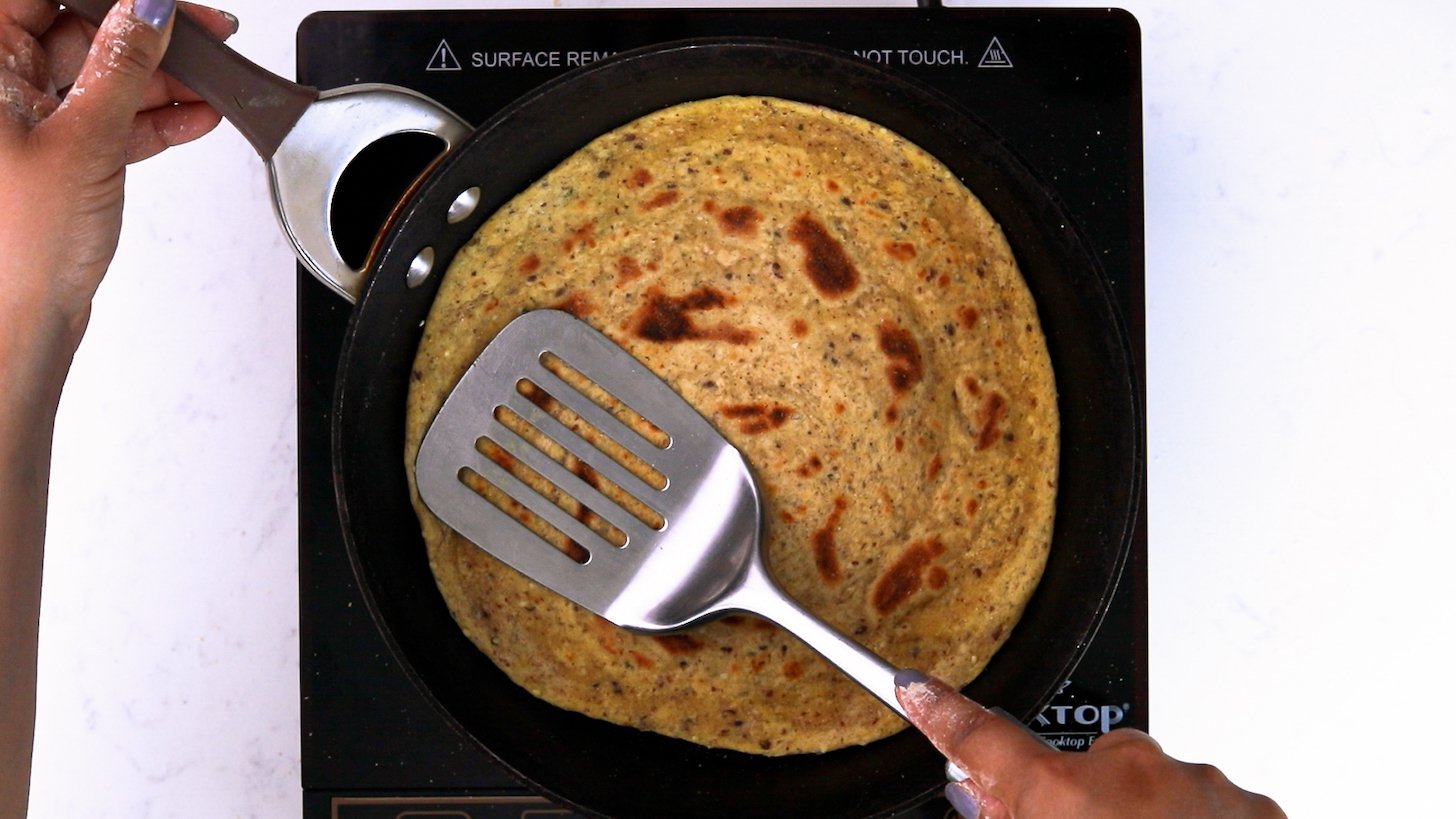 A hand holding a metal spatula pressing flatbread (paratha) in a pan.