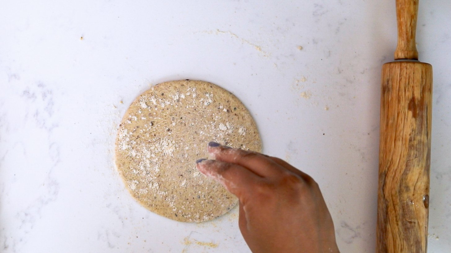A hand sprinkling flour on a circular rolled-out dough with a rolling pin close by.