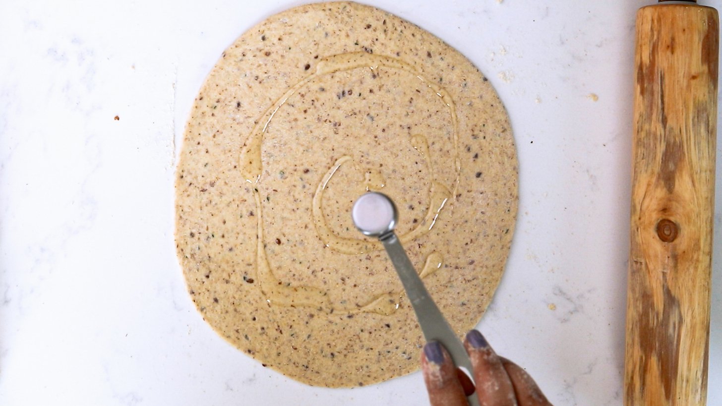 Hand holding a spoon drizzling oil on rolled-out circular dough with a rolling pin close by.