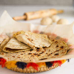 A decorative basket of flatbread (paratha) - with one ripped in half stacked on top of one another. There is a rolling pin and dough in the background.
