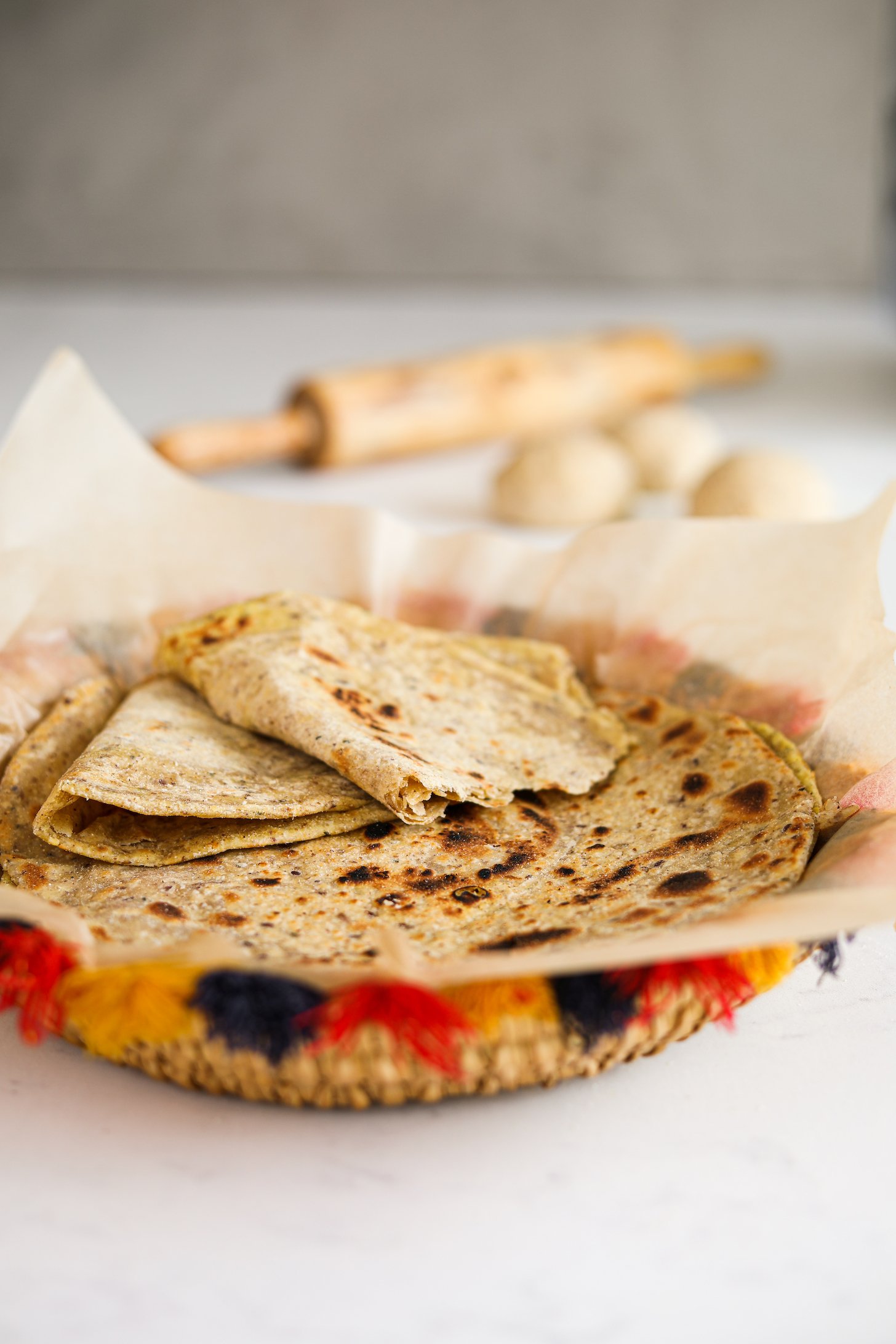 A decorative basket of flatbread (paratha) - with one ripped in half stacked on top of one another. There is a rolling pin and dough in the background.