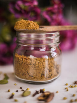 A perspective image of a wooden heaped full spoon of brown powdered spice balancing on the rim of a mason jar with a bouquet of flowers in the background.