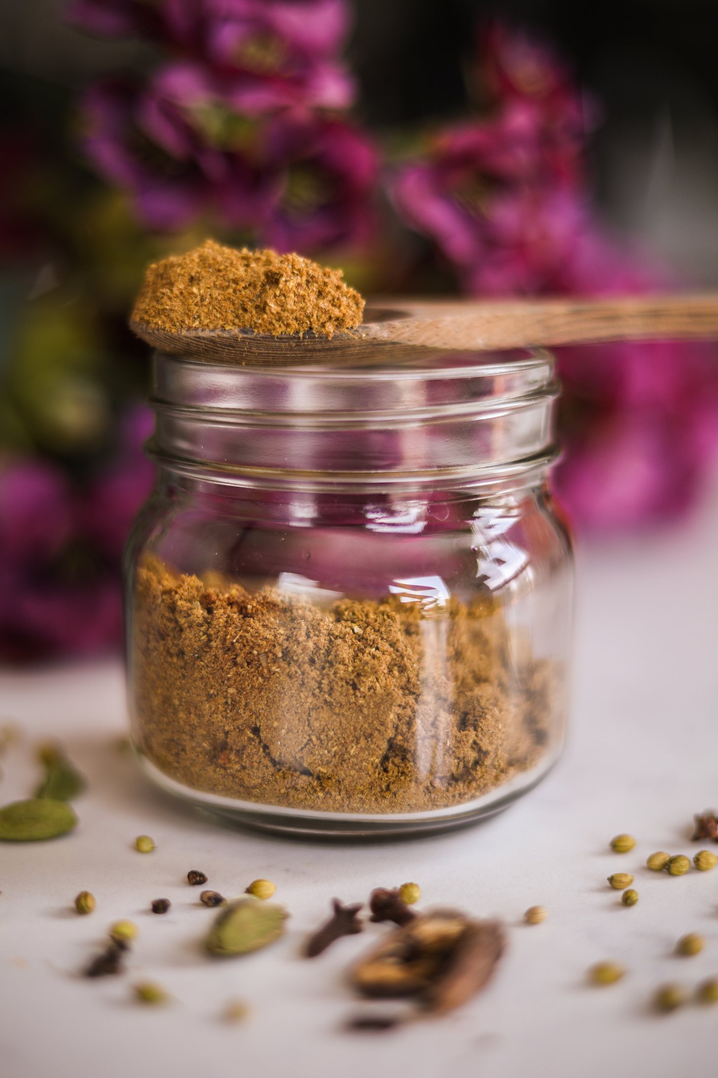 A perspective image of a wooden heaped full spoon of brown powdered spice balancing on the rim of a mason jar with a bouquet of flowers in the background.
