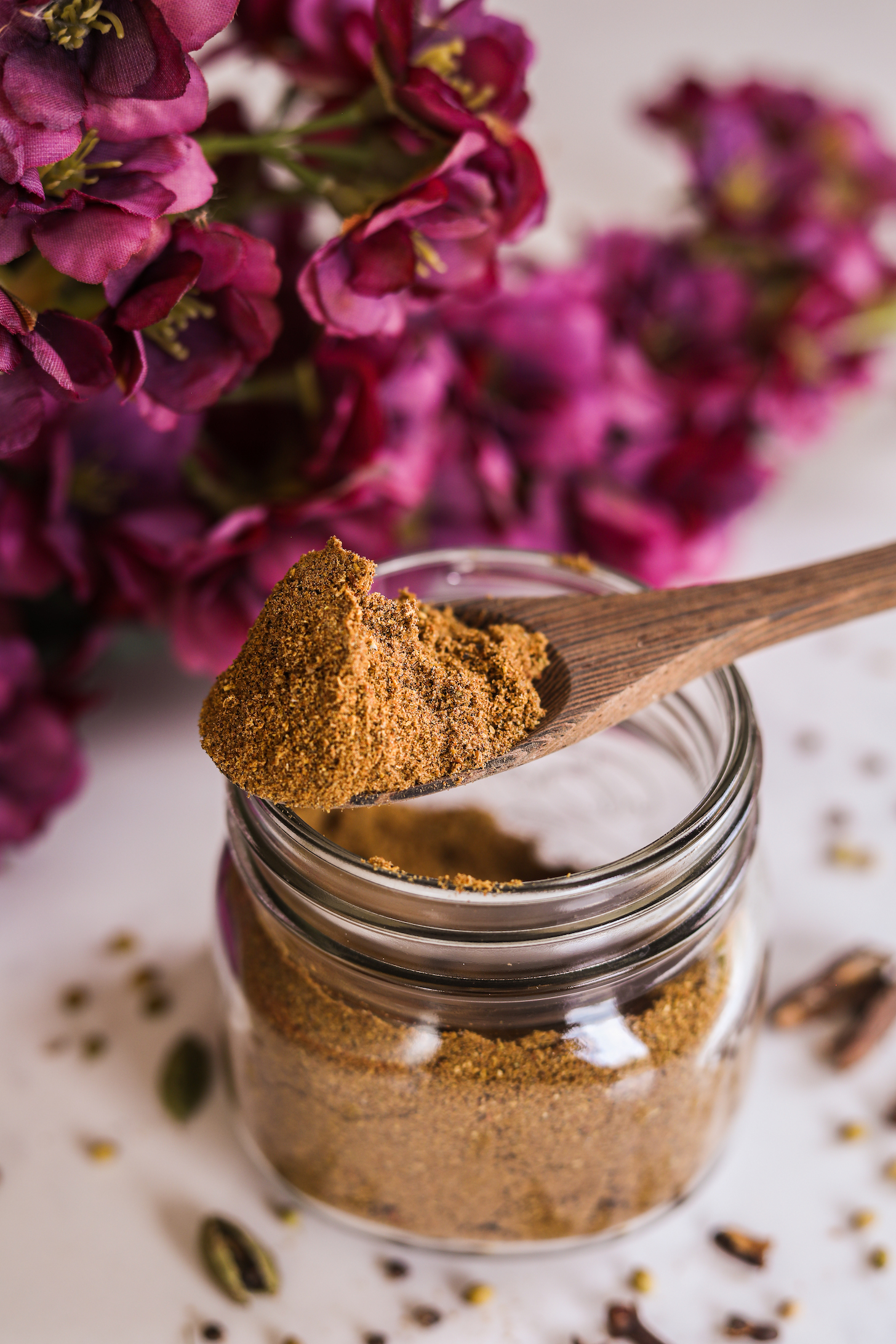 A heaped spoon of brown spice balancing on a mason jar with pink flowers in the background.