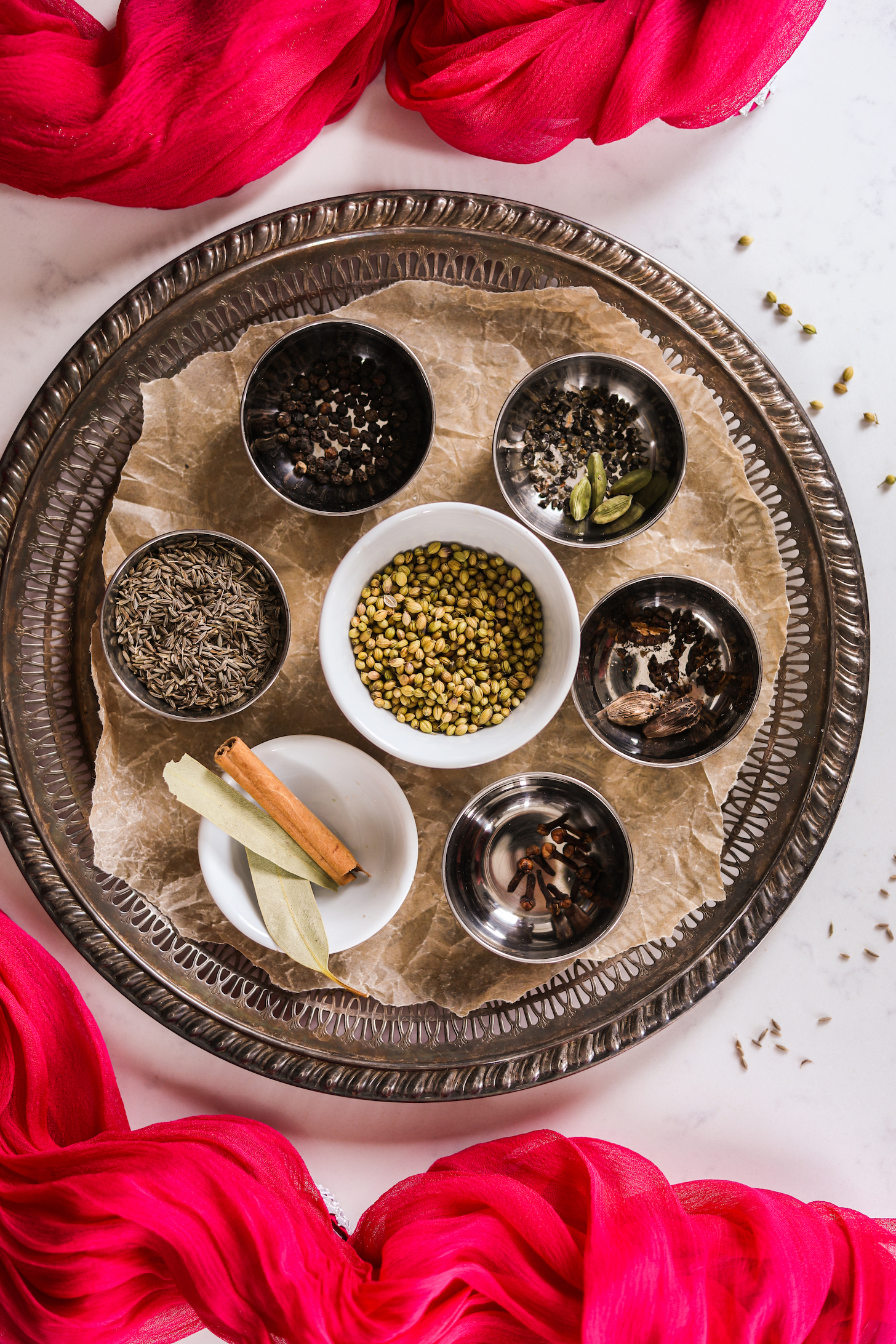 A round tray with ramekins of whole South Asian spices like cloves, cardamoms, coriander seeds and more.