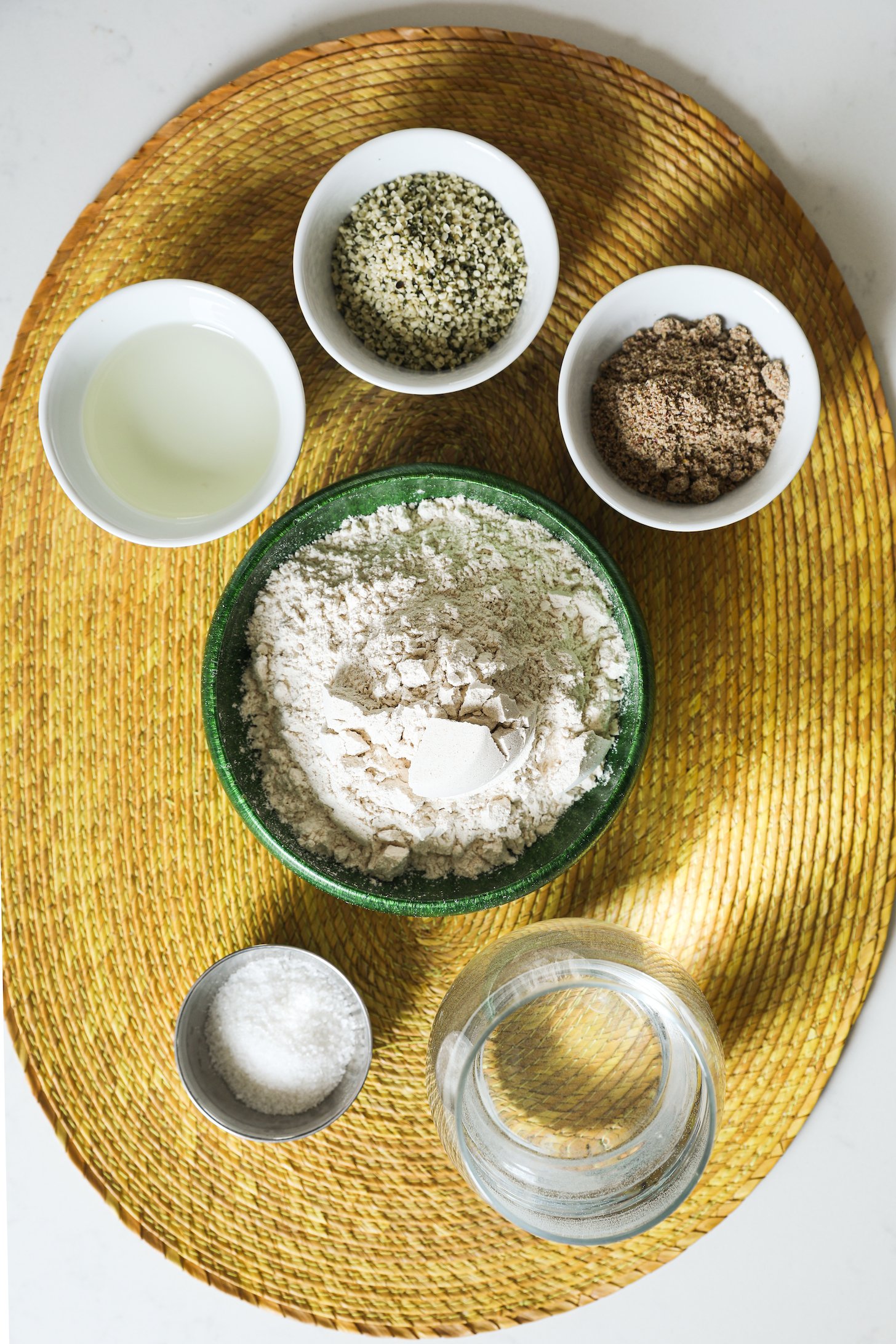 Food ingredients (flour, seeds, water, salt and oil) in bowls and ramekins on a yellow straw mat.