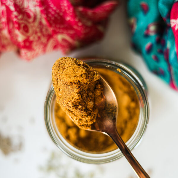 Top view of a gold spoon of yellow spice powder sitting on a jar with a pink and green scarf in the background.