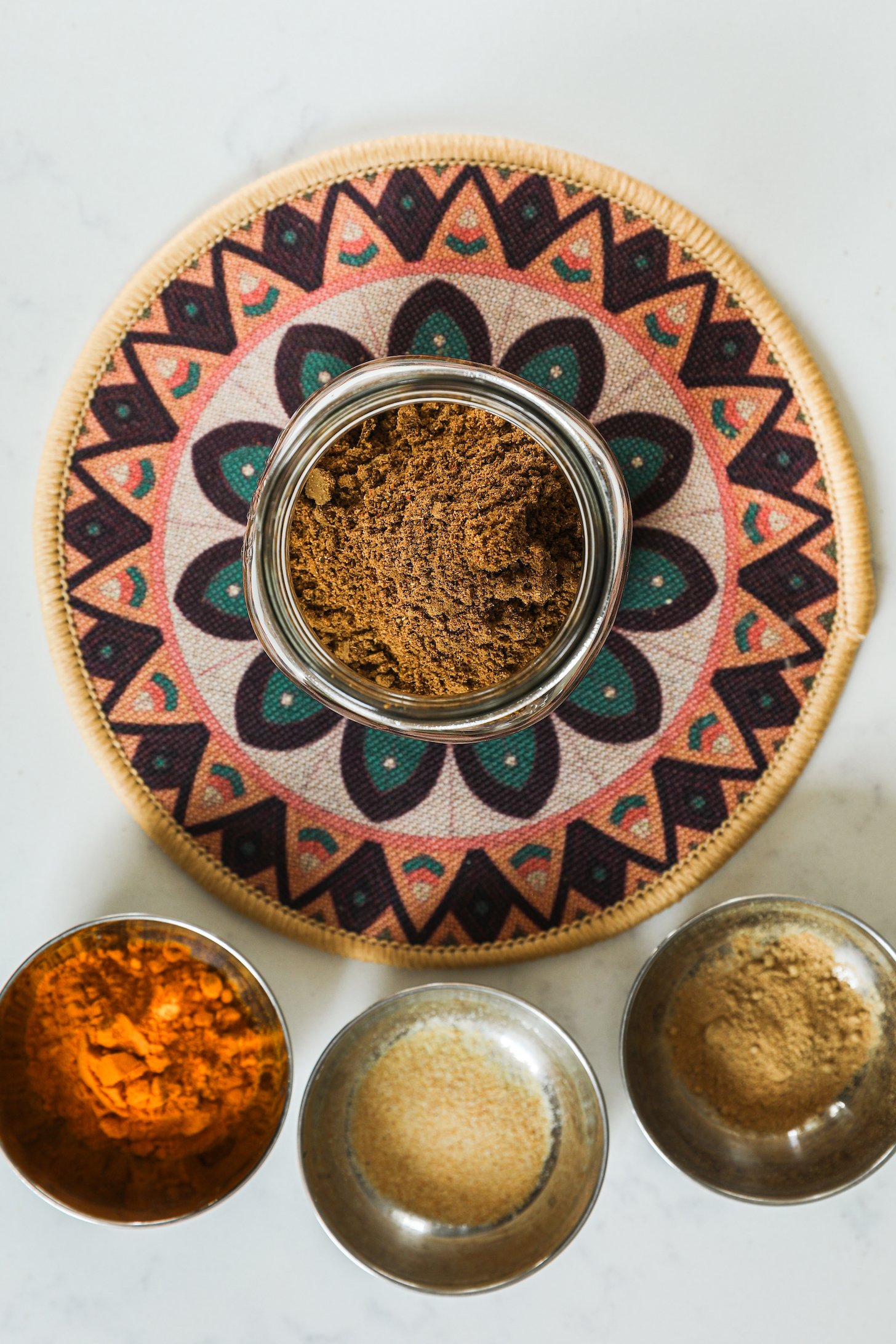 Top view of a jar of ground brown spices on a patterned placemat with 3 ramekins of ground spices arranged close by.