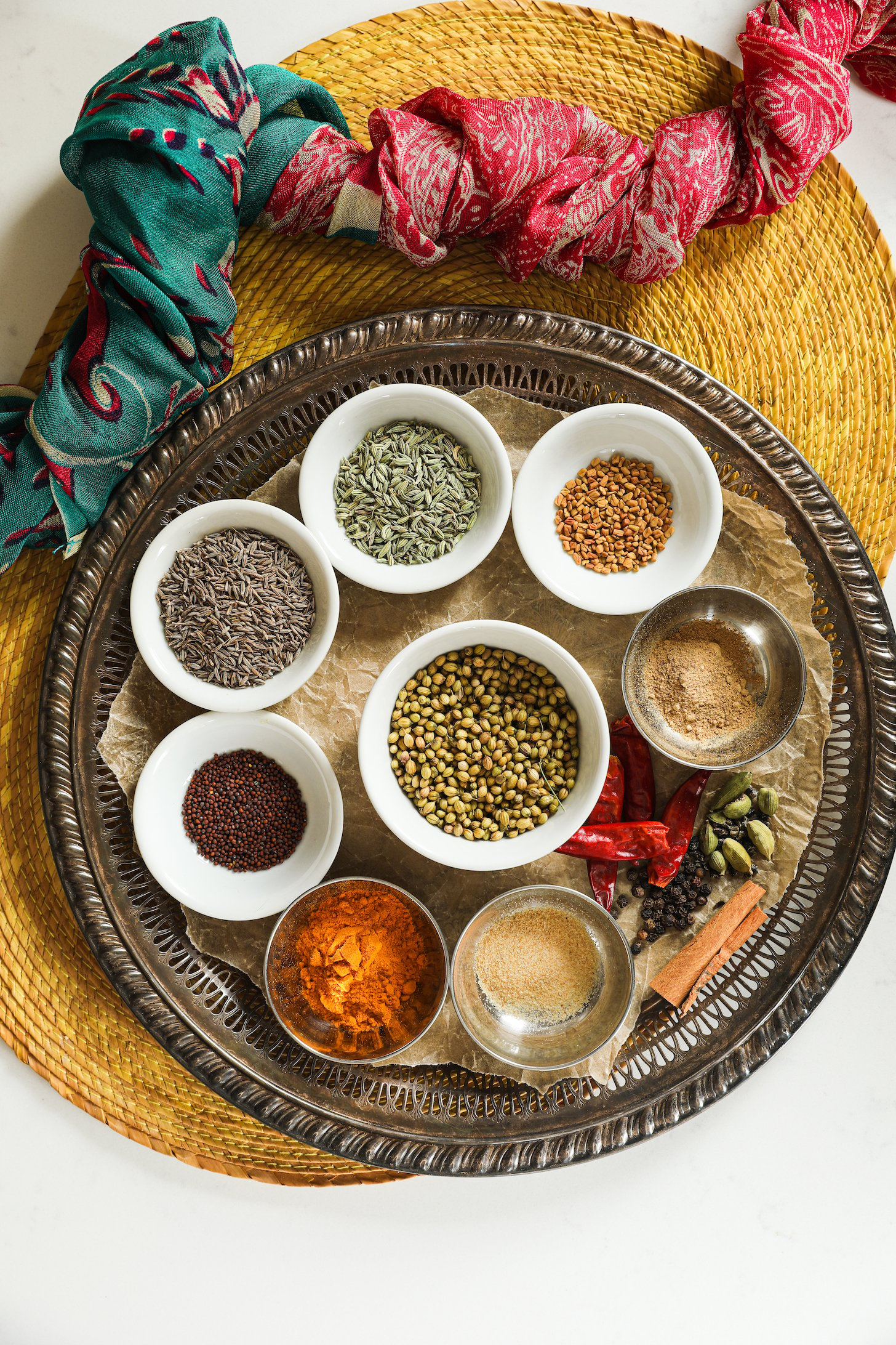 A collection of whole South Asian whole spices arranged beautifully in a a traditional round tray with a scarf on one side.