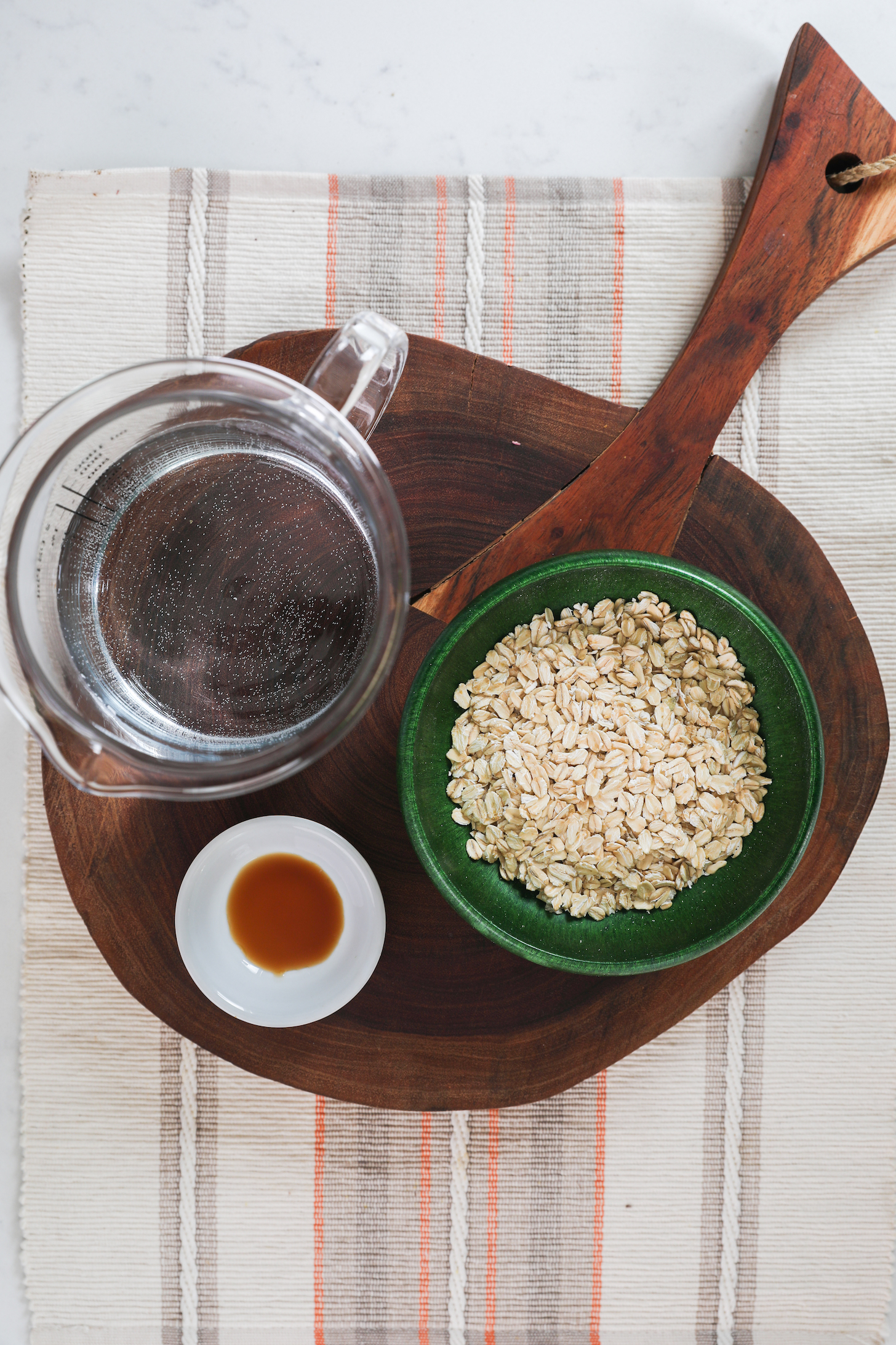 A bowl of rolled oats, a ramekin with vanilla and a jog of water styled on a wooden board.