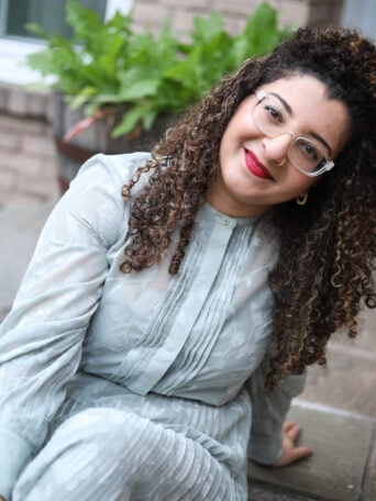 A curly-haired woman sitting on the ground smiling and posing.