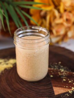 Perspective view of a mason jar of yellow dressing with plant and flowers in the background.