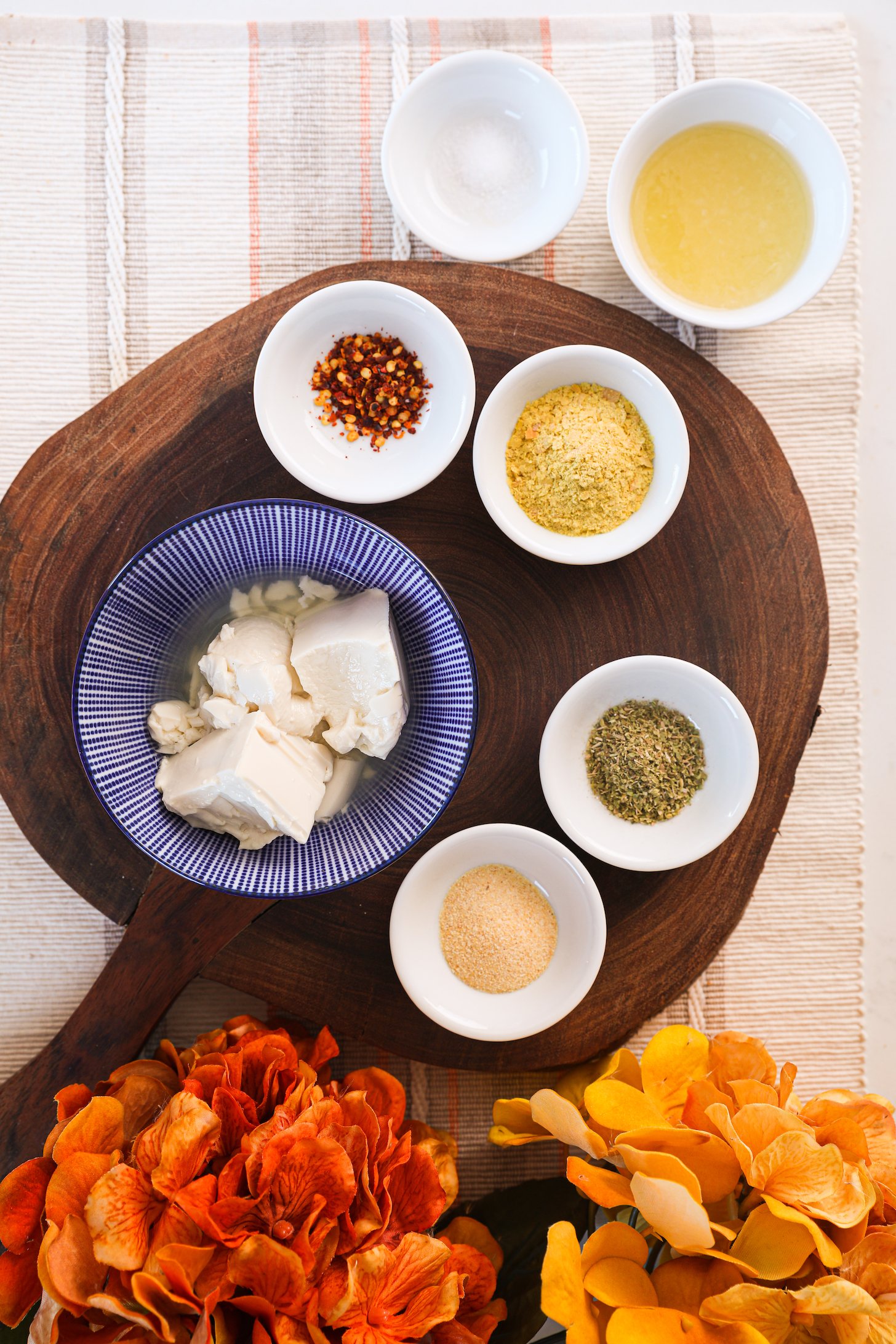 A selection of food ingredients arranged on a wooden board including soft tofu, yeast and spices with bright yellow flowers.