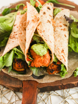 Perspective view of three chicken tikka wraps piled on top of fresh greens on a wooden board.