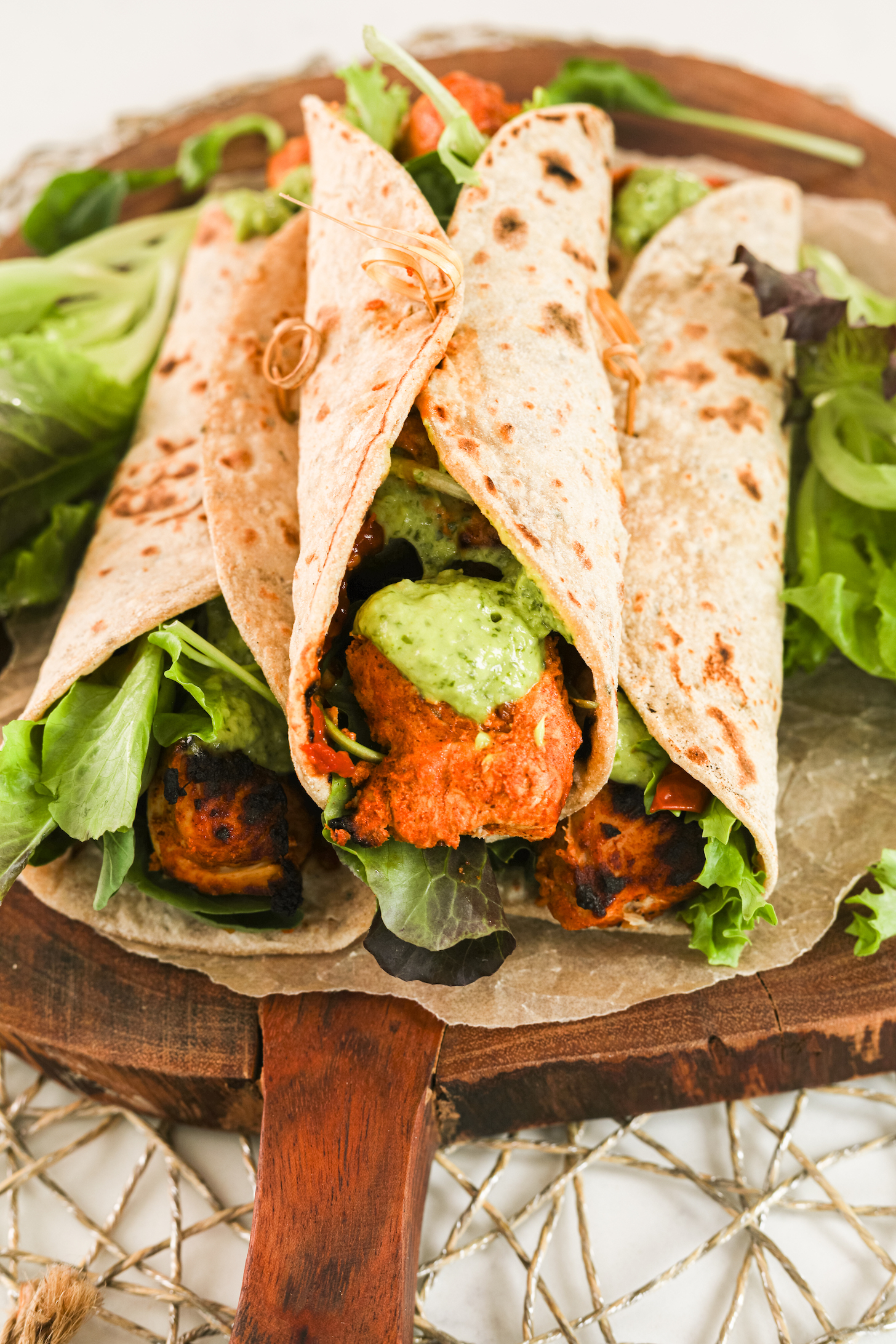 Perspective view of three chicken tikka wraps piled on top of fresh greens on a wooden board.