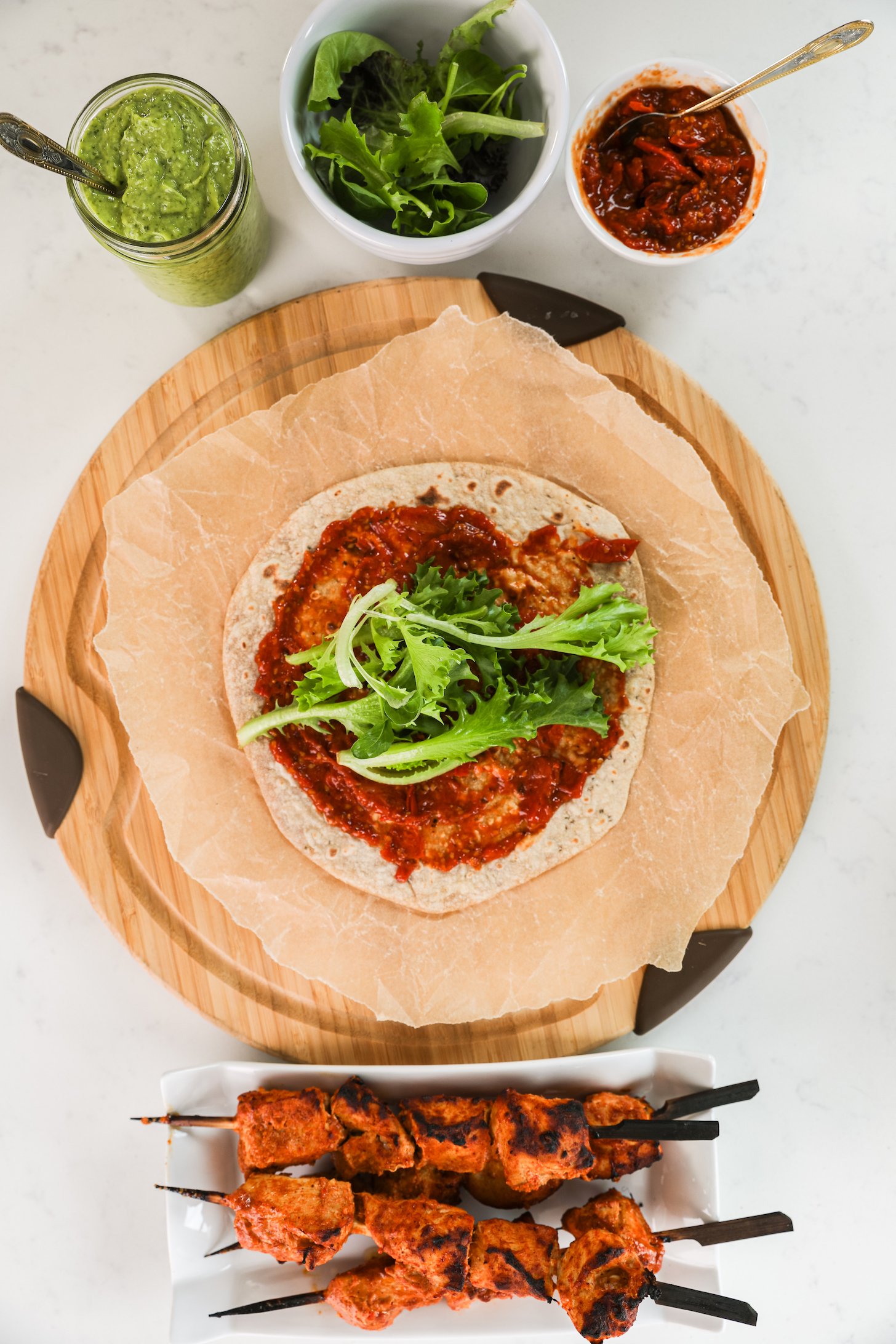 A roti spread with tomato sauce and greens with tandoori chicken skewers, sauces and salad greens nearby.