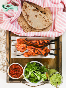 A selection of food ingredients like grilled tandoori chicken skewers, greens, sauces and roti.